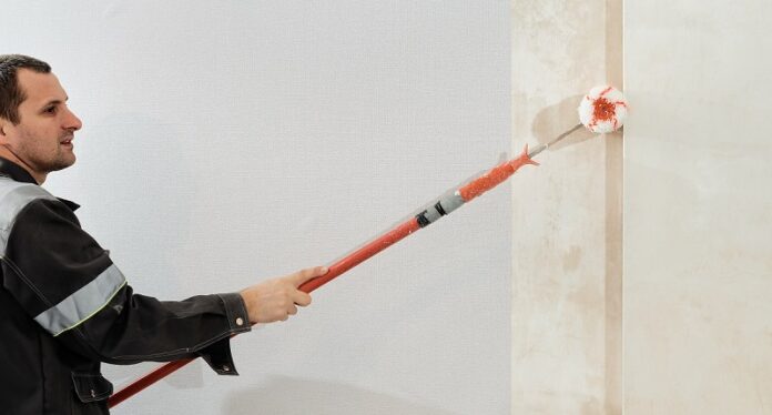 Man worker applies glue with a paint roller on the walls before wallpapering or primer on the walls, selective focus. Apartment renovation and restoration idea indoors.